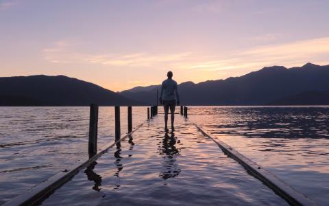 Silhouette of a person from behind standing at the end of a dock looking out at a lake. The sun is rising behind the mountains.